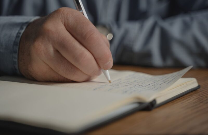 A hand writing with a pen in an open notebook on a wooden desk