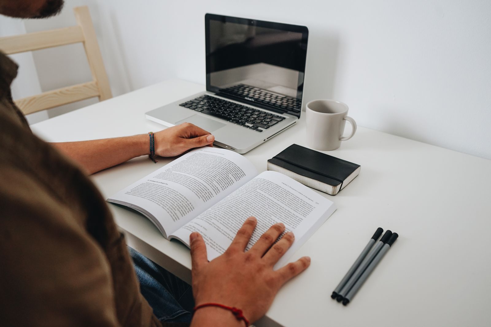 A person reading at a tidy desk with an open laptop, a book and a coffee mug