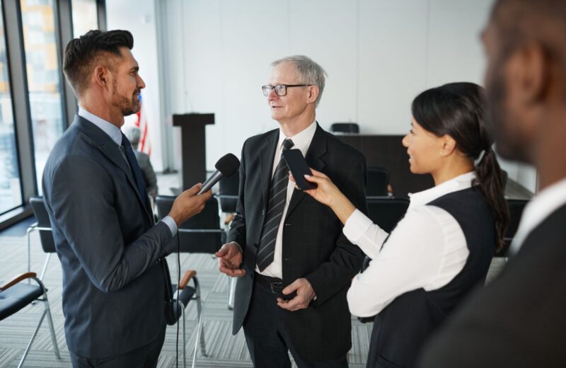 Journalists holding microphones towards a man in a suit during an interview