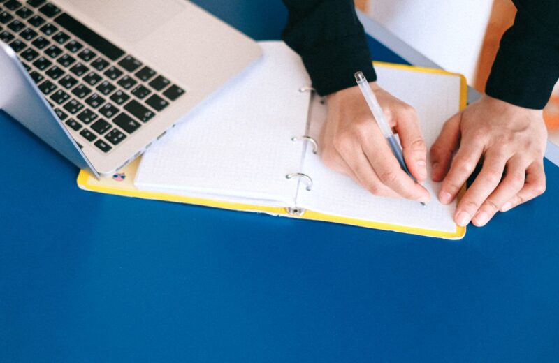 Hands writing in a notebook next to a laptop on a blue table
