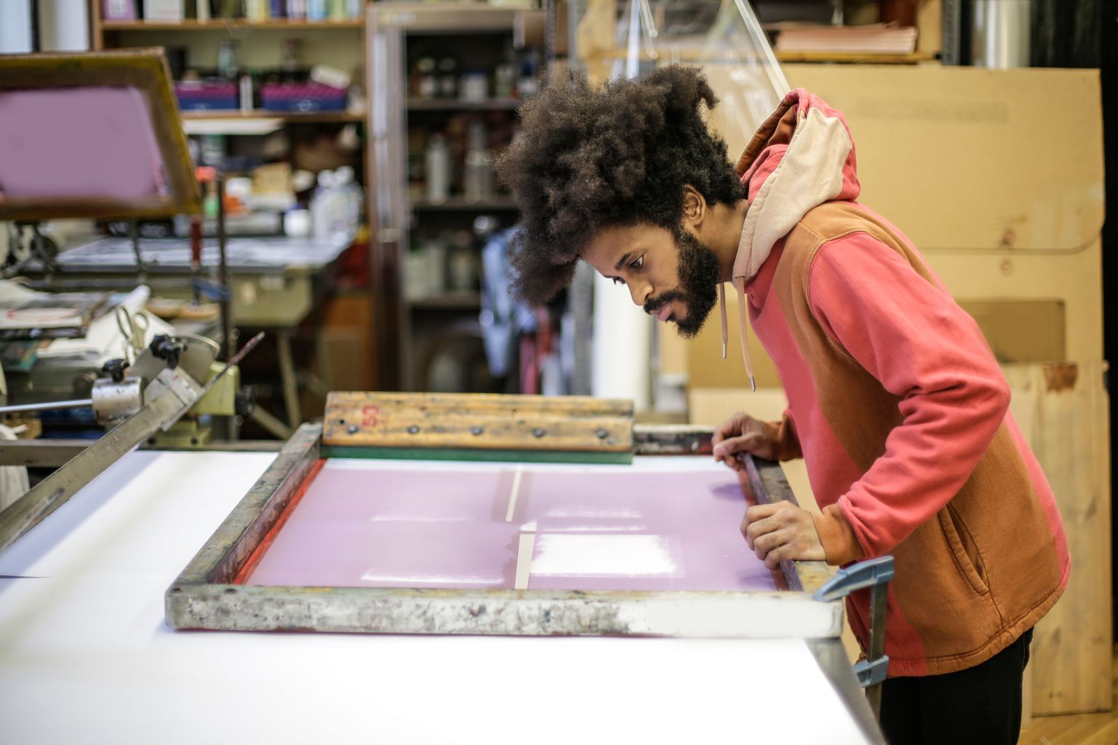 A printmaker leaning over a pink screen-printing frame on a long print table