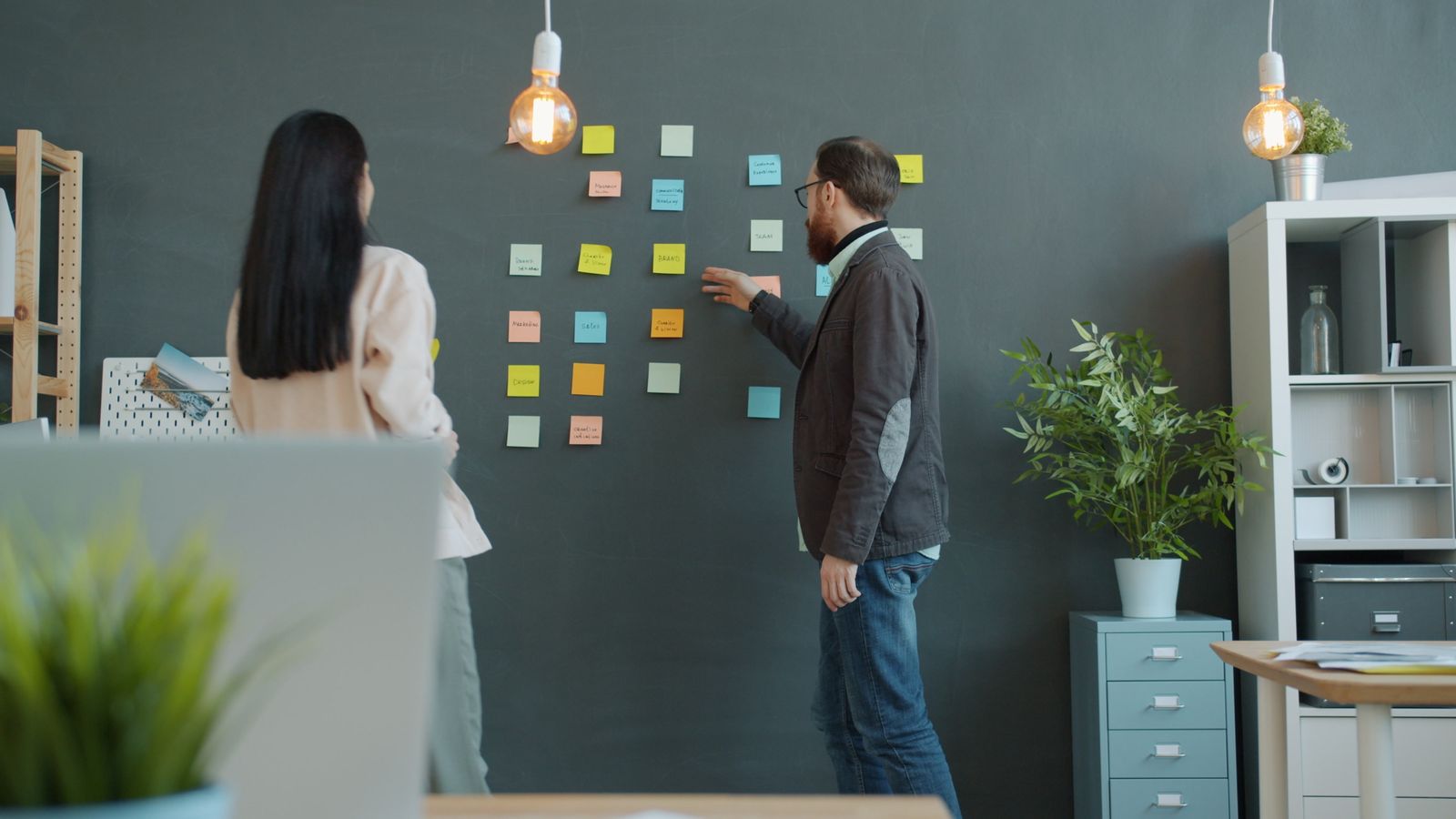 Two people from behind, moving sticky notes on a dark wall in a bright studio