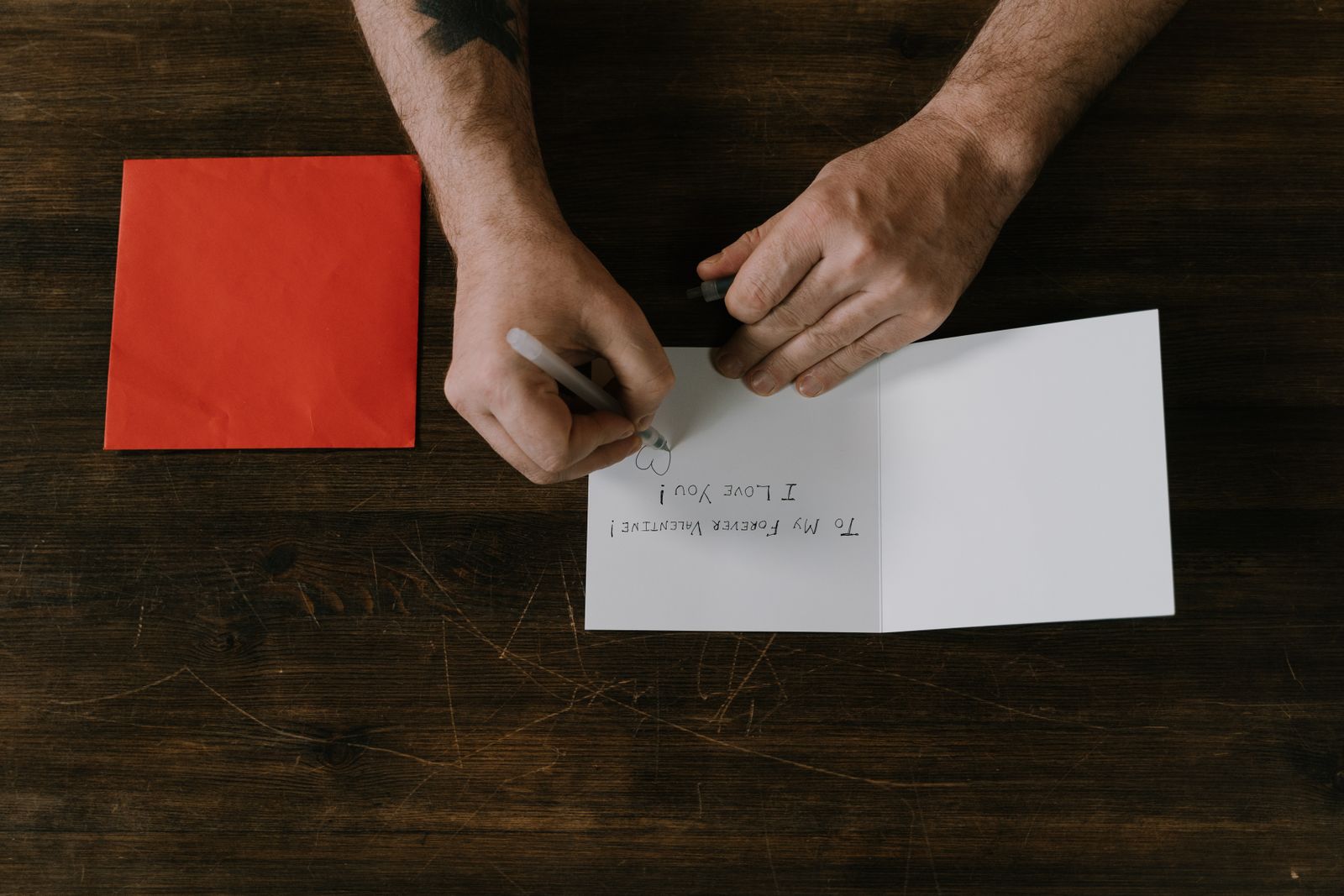 Seen from above, a man's hands writing on a white card at a dark wooden table, a red envelope beside it