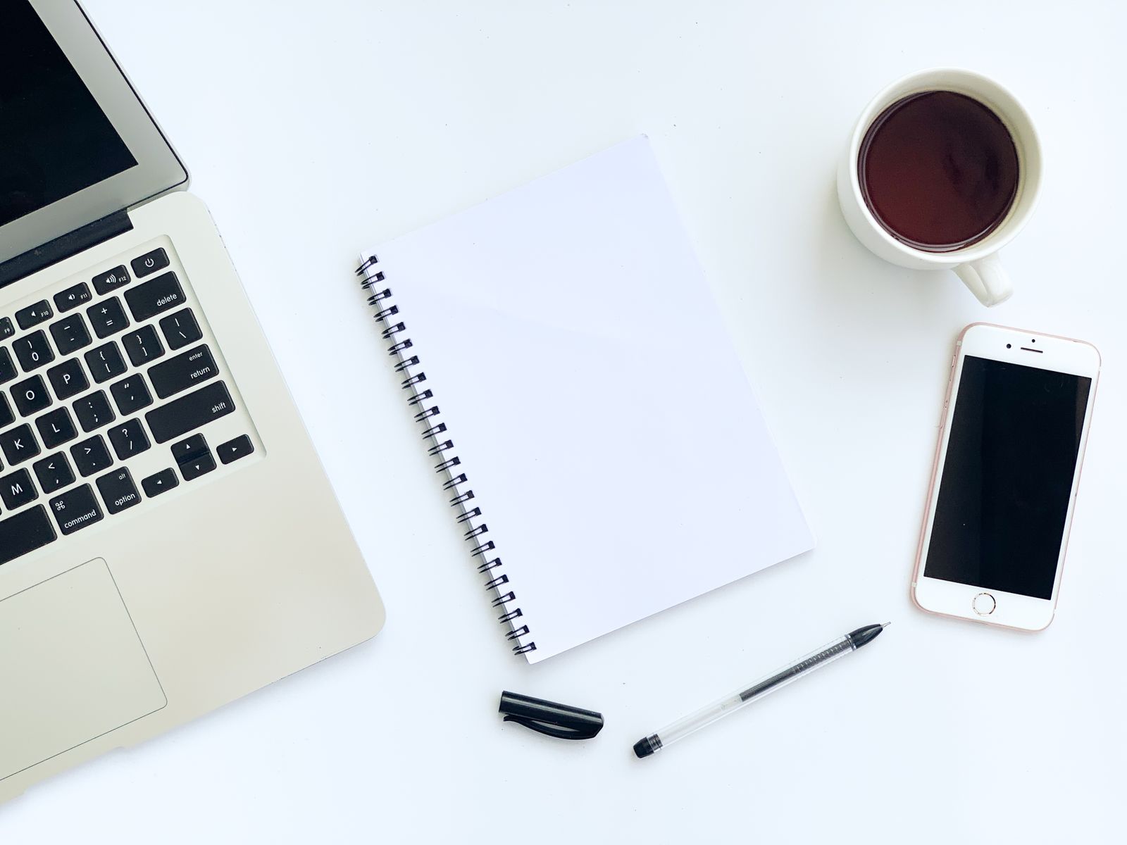 A tidy white desk from above with a laptop, a spiral notebook, a pen, a phone and a cup of tea