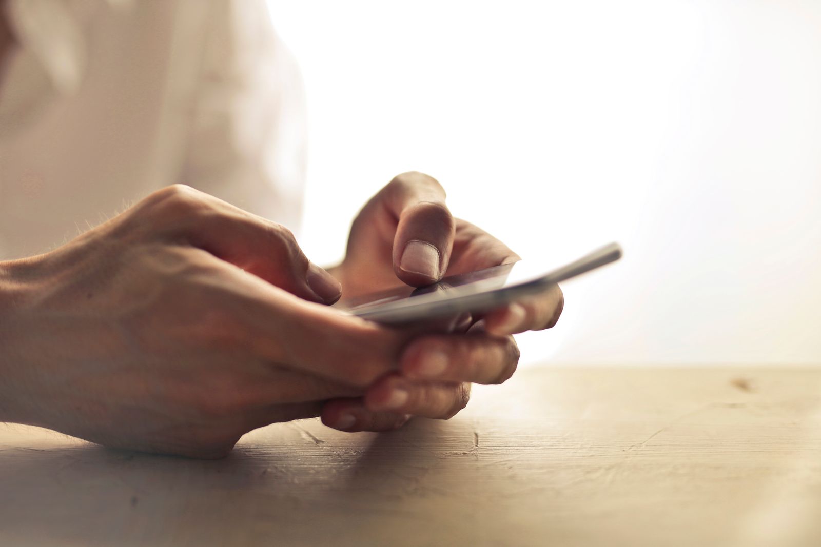 Two hands holding a smartphone over a pale wooden table in soft window light