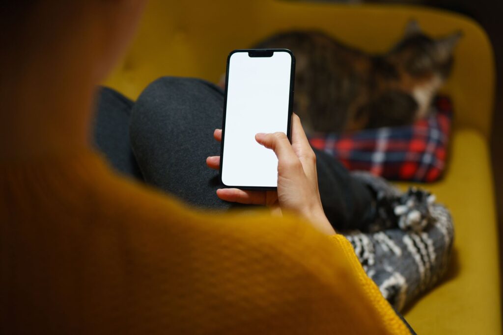 A person on a yellow sofa holding a phone with a blank screen, a cat asleep beside them