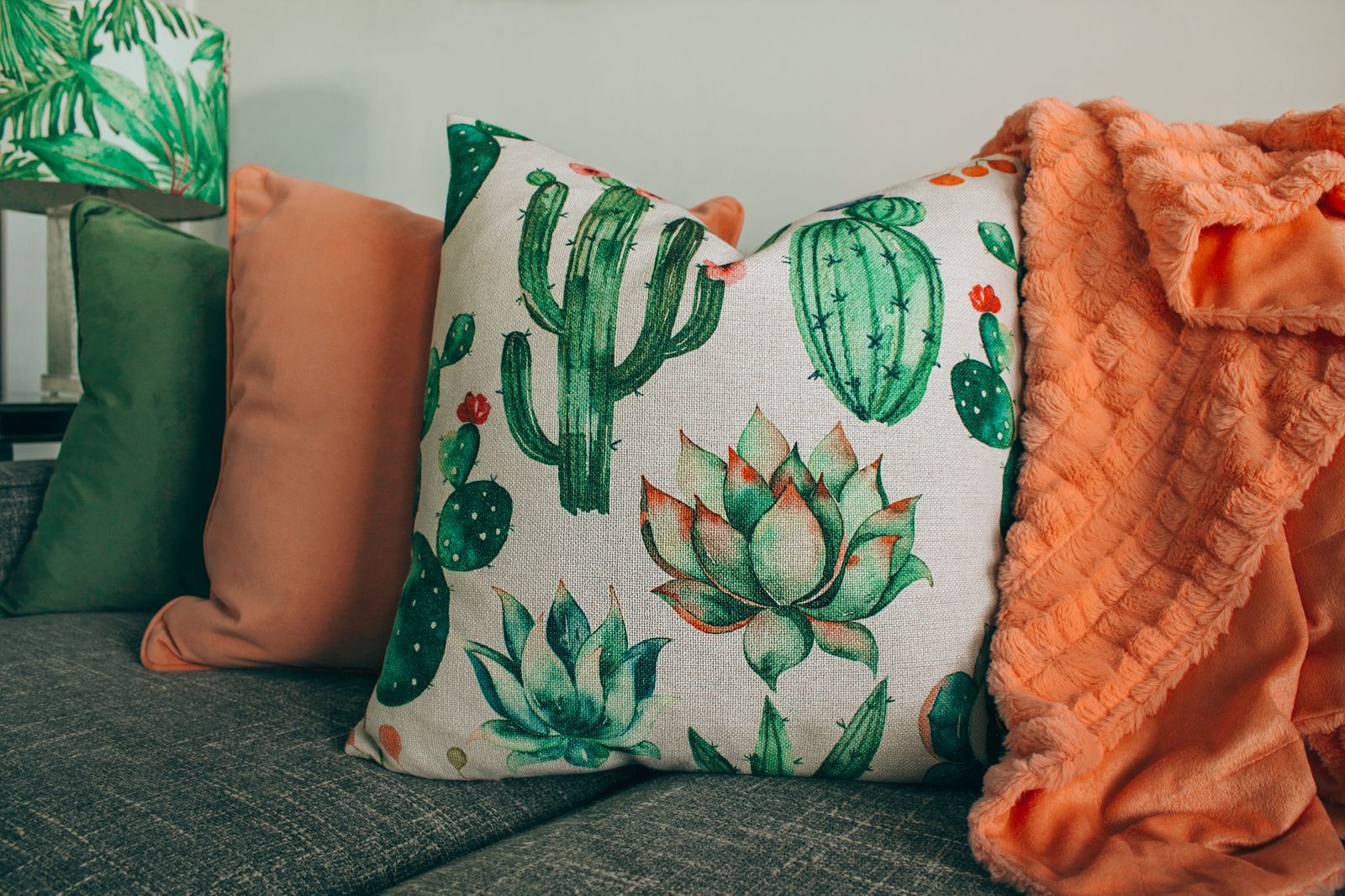 Cushions printed with green cacti, a plain pink cushion and an orange knitted throw on a grey sofa