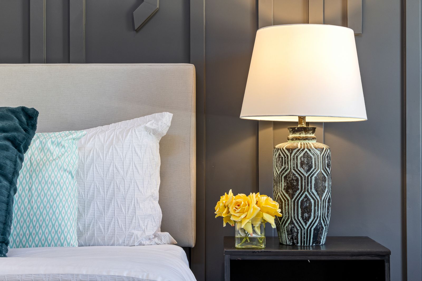 A patterned ceramic table lamp with a white shade on a dark bedside table, yellow flowers beside it