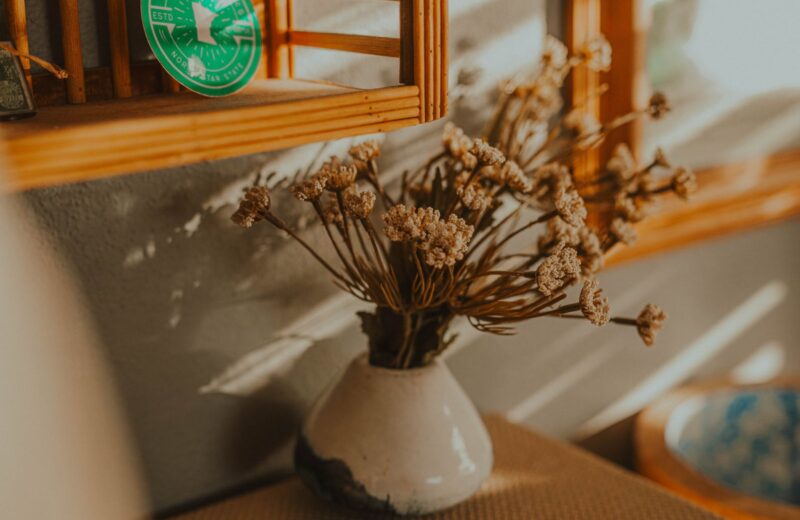 A white vase of dried flowers on a wooden shelf in afternoon sun