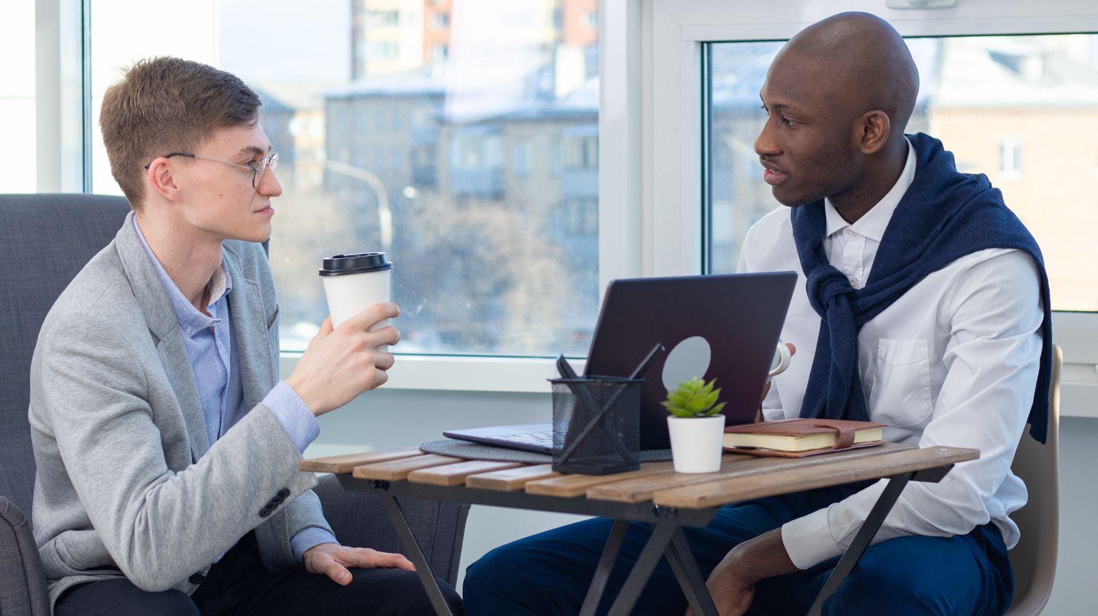 Two people talking over laptops and coffee at a table by the window