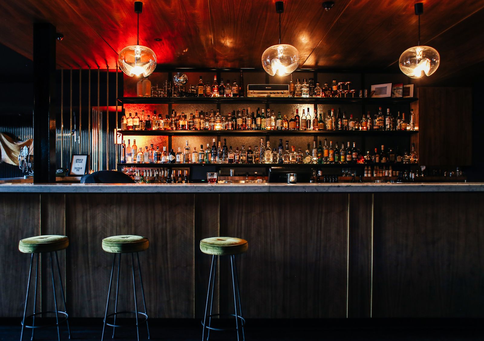 A dark wooden bar counter with three stools and lit shelves of bottles