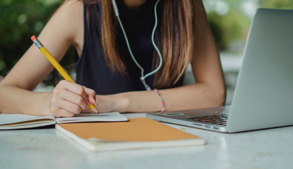 A person writing notes in a notebook beside a laptop at a table