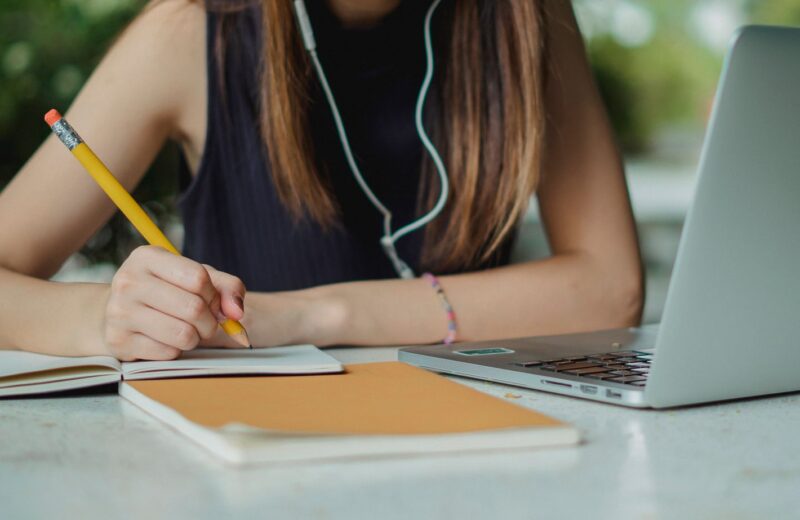 A person writing notes in a notebook beside a laptop at a table