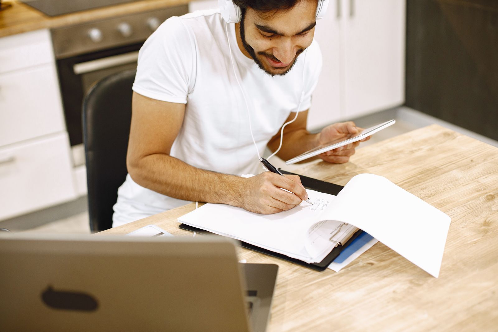 A man taking notes from a tablet and a laptop at his kitchen table