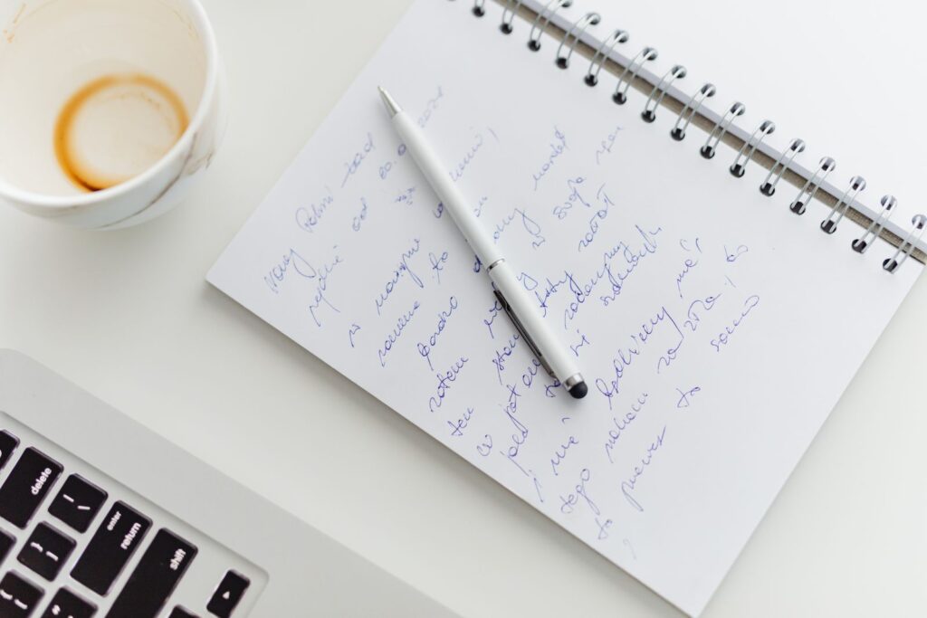 A notebook of handwritten notes and a pen beside a laptop and a coffee