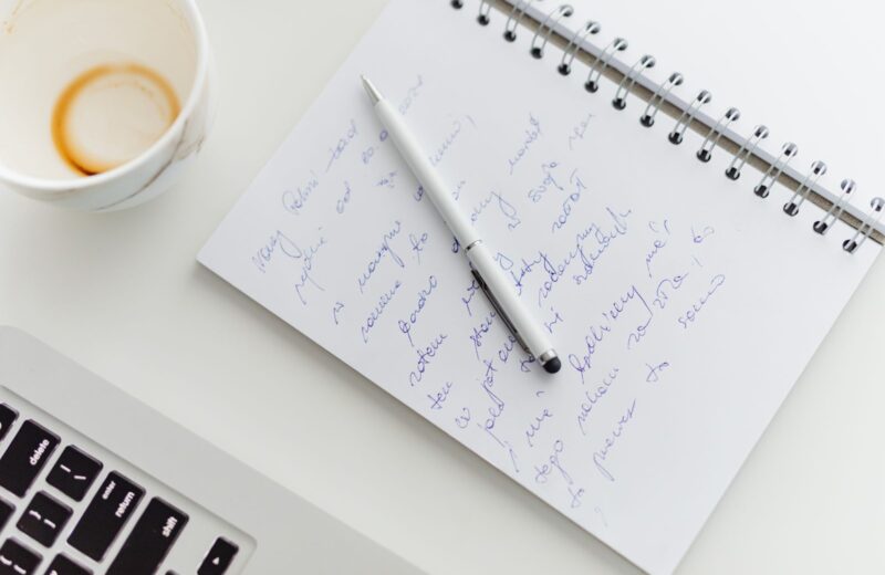 A notebook of handwritten notes and a pen beside a laptop and a coffee