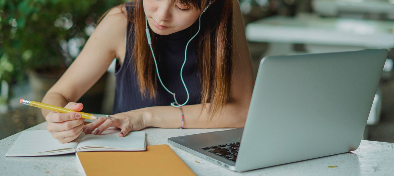 A woman with earphones writing notes beside her laptop at a table