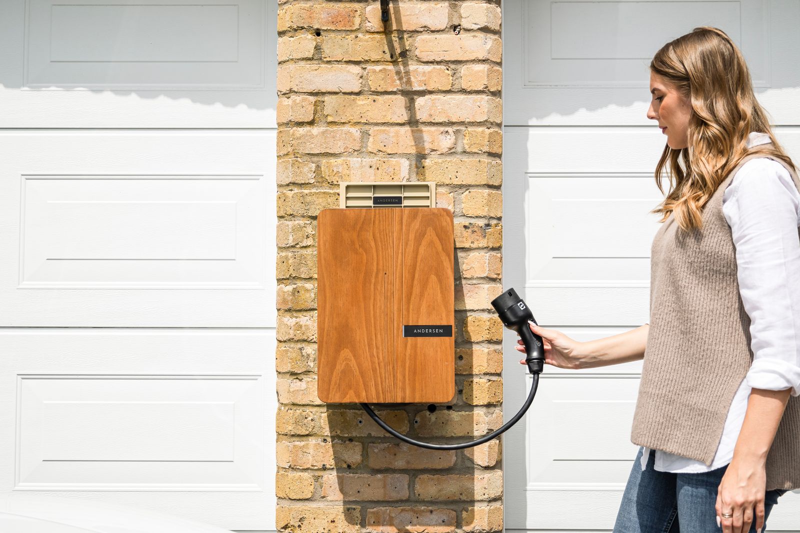 A woman taking the charging cable from a home EV charger on a brick garage wall