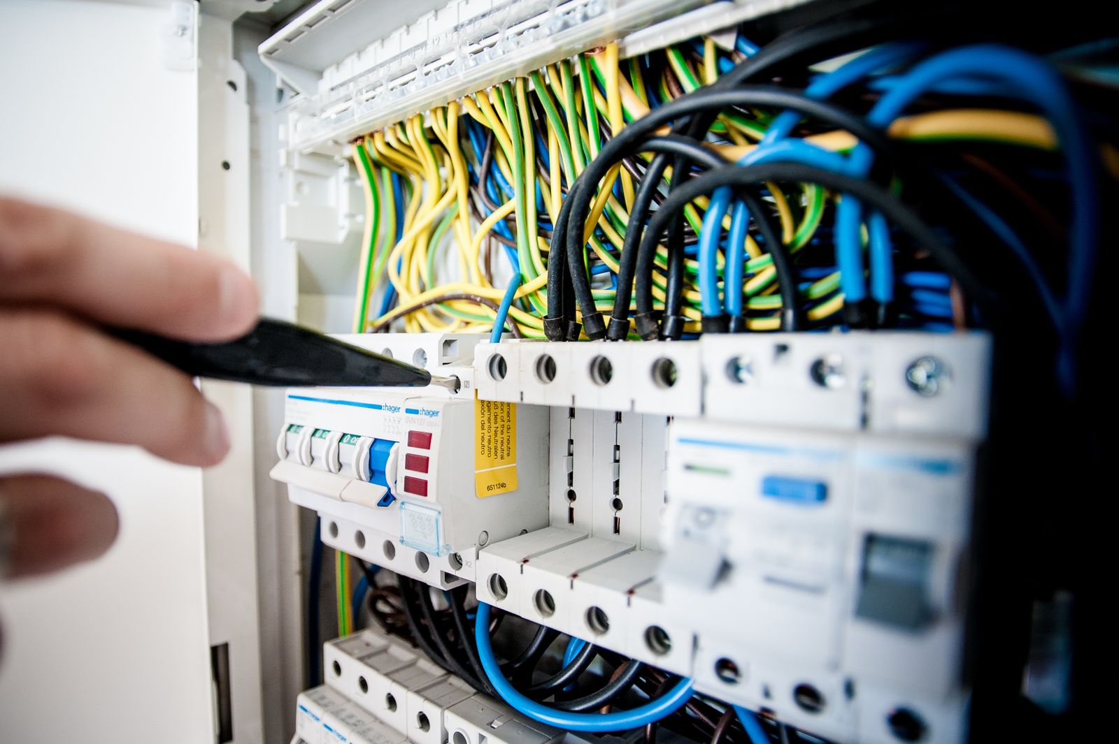 A hand pointing a screwdriver at breakers in an open panel with colour-coded wires