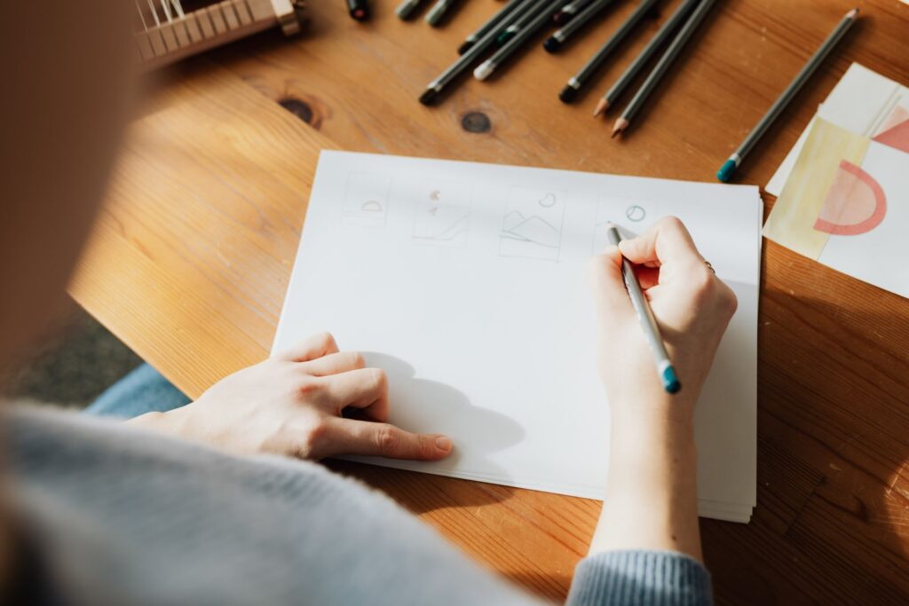 Seen from above, a hand drawing on a blank sheet beside a row of pens