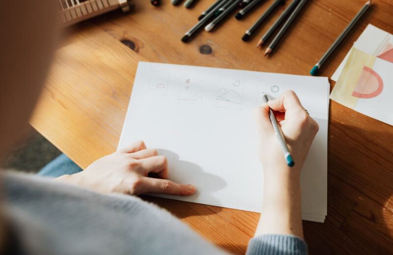 Seen from above, a hand drawing on a blank sheet beside a row of pens