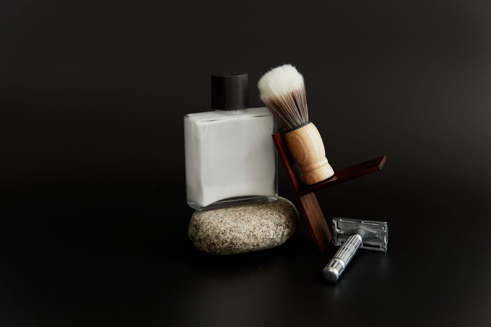 A shaving brush, a safety razor and a white bottle on a pebble against a black background