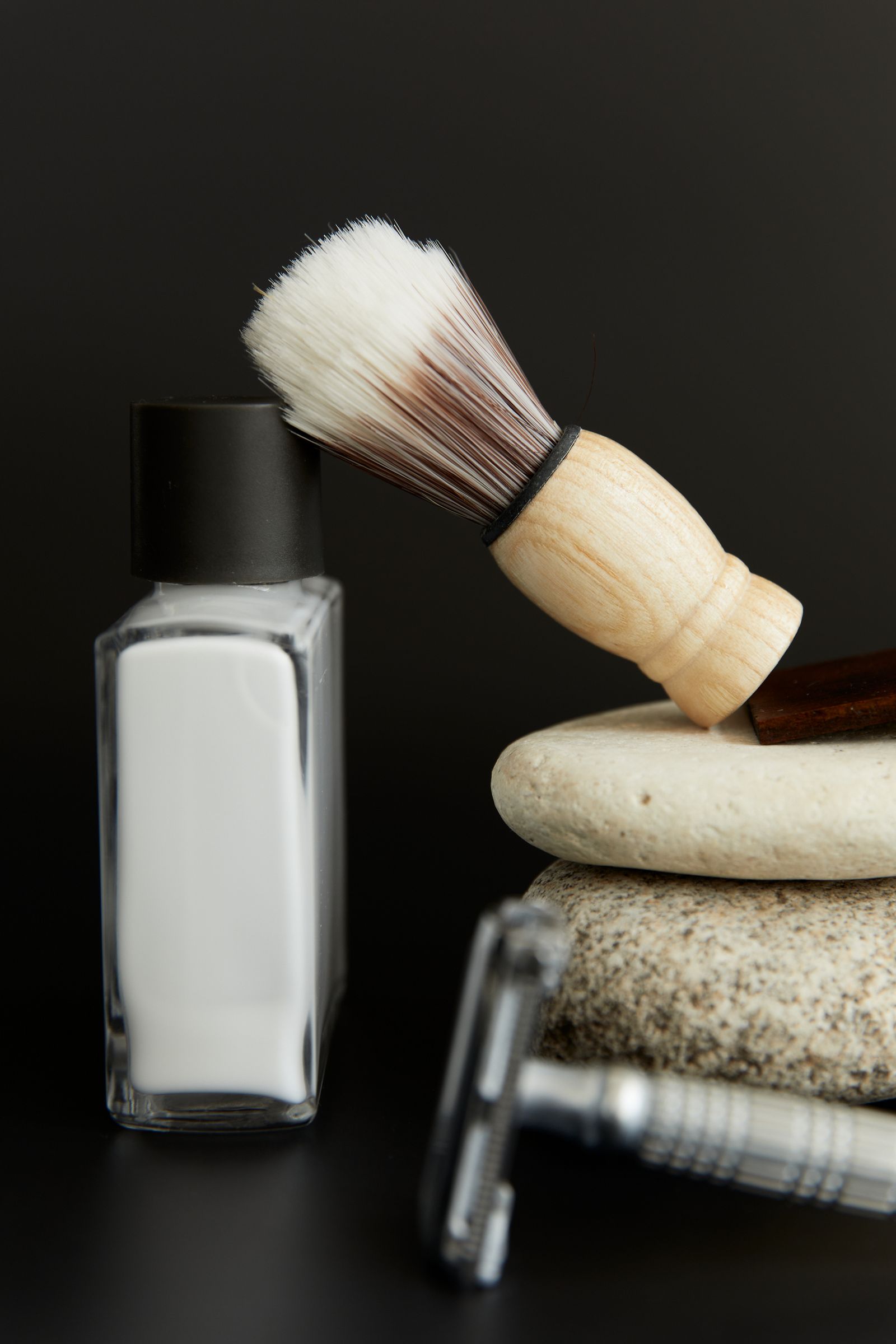 A shaving brush with a pale wooden handle resting on a stone beside a white bottle