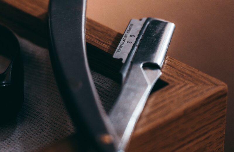 A straight razor with a dark handle resting open on the edge of a wooden box in warm light