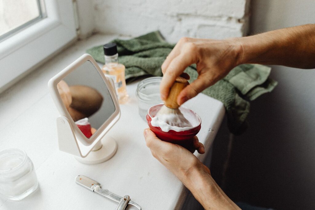 Hands working up shaving cream with a brush in a red bowl by a bathroom window