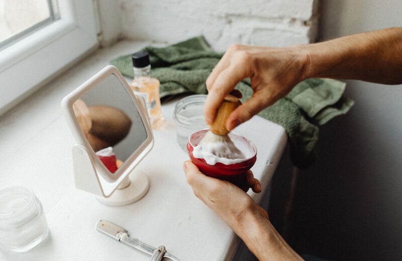 Hands working up shaving cream with a brush in a red bowl by a bathroom window