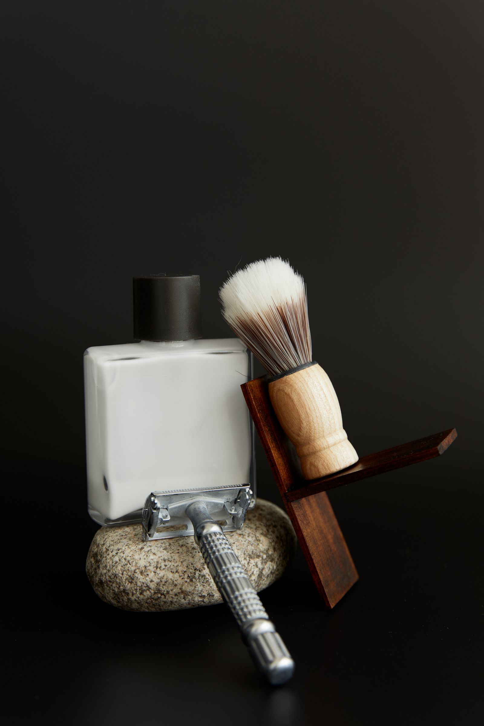 A white bottle and a shaving brush on a wooden stand, with a razor, on a black background