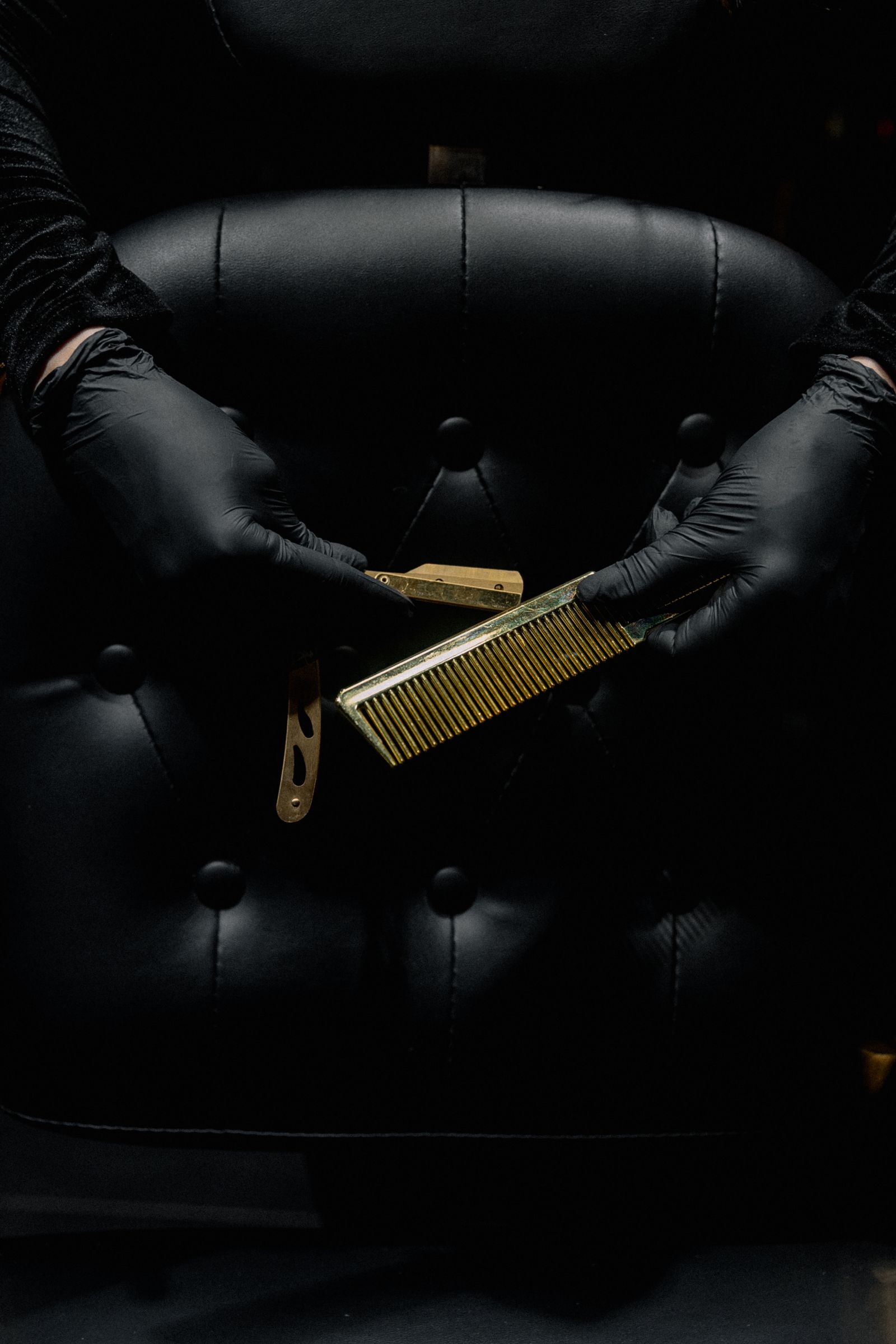 A barber's gloved hands holding a comb and a razor in front of a black leather chair