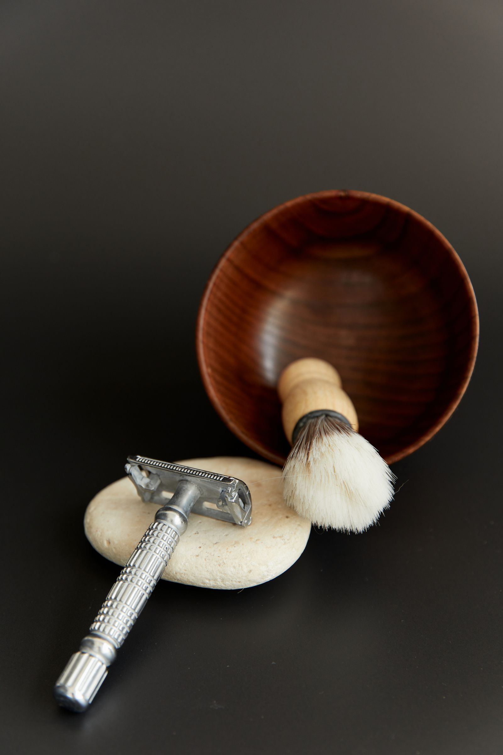 A wooden lather bowl with a shaving brush in it and a safety razor resting on a pale stone