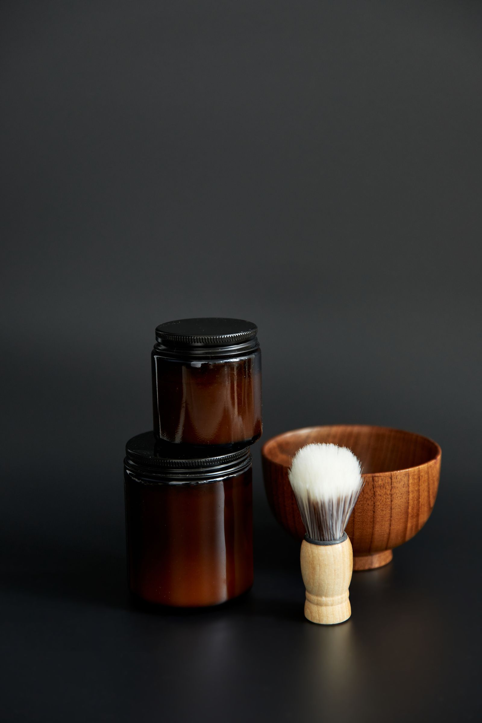 Two brown glass jars of shaving soap and a wooden bowl with a white brush on a dark grey background