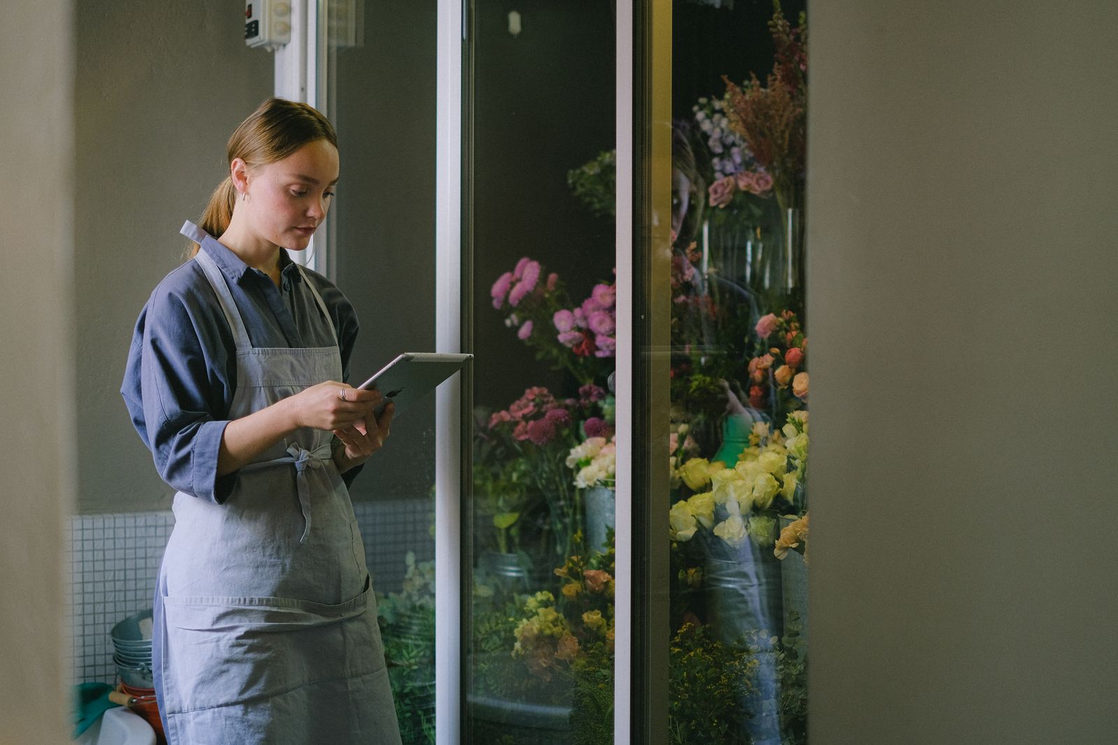 A florist in an apron reading a tablet beside a flower fridge