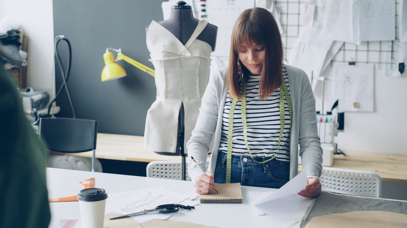 A designer sketching at a cutting table next to a dress form with a toile pinned on it