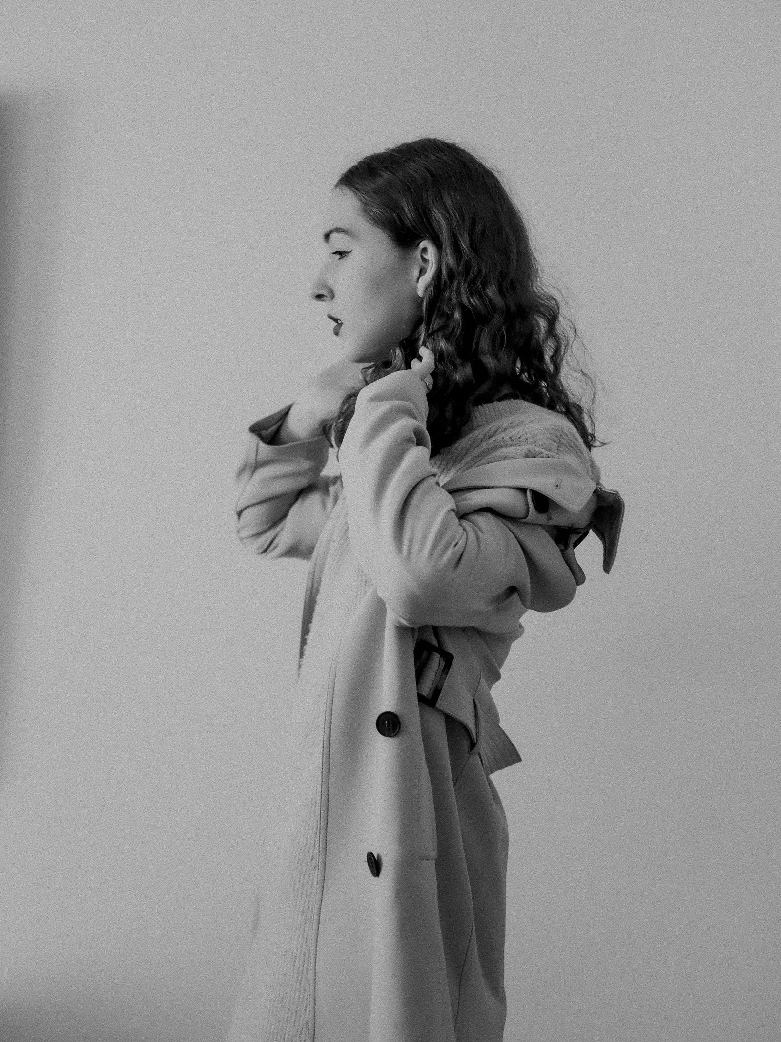 Black and white photo of a woman in a pale wool coat turning up its collar against a plain studio wall