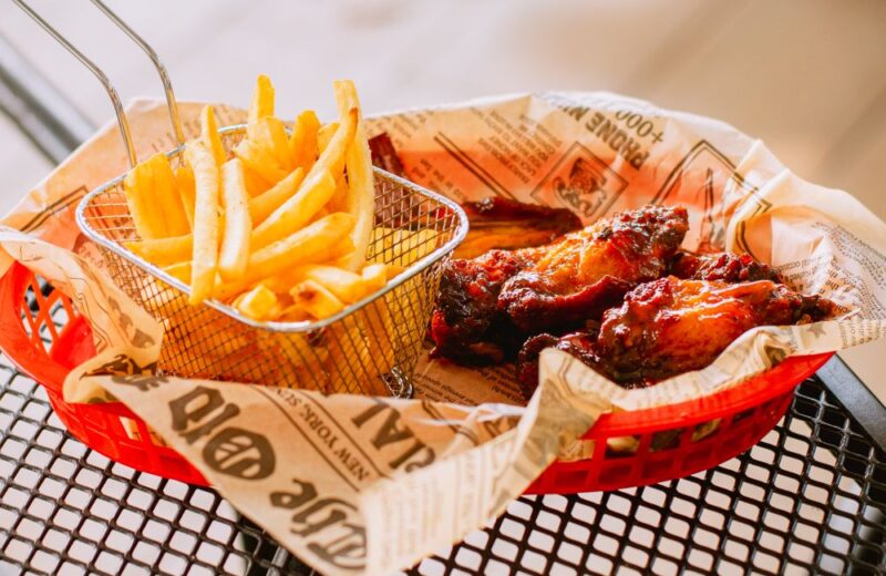 A wire basket of fries beside glazed chicken wings on printed paper in a red tray