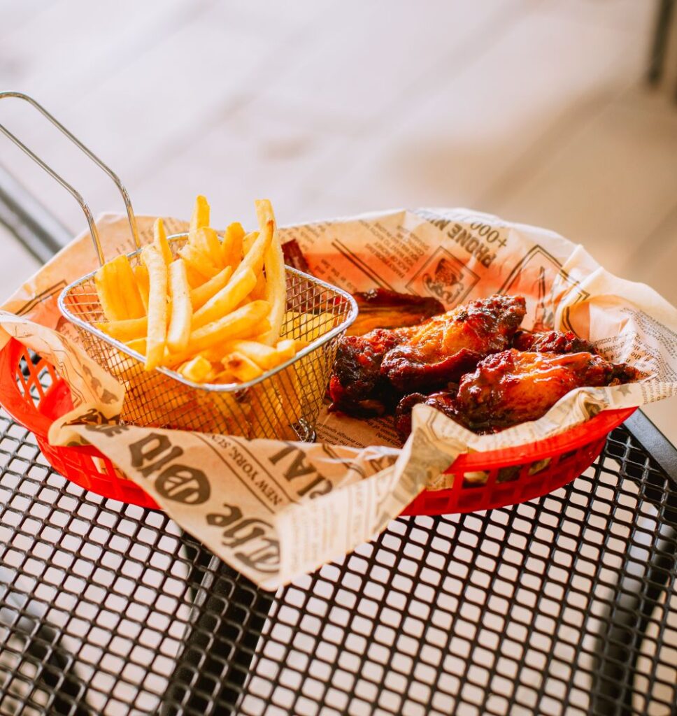 A wire basket of fries beside glazed chicken wings on printed paper in a red tray