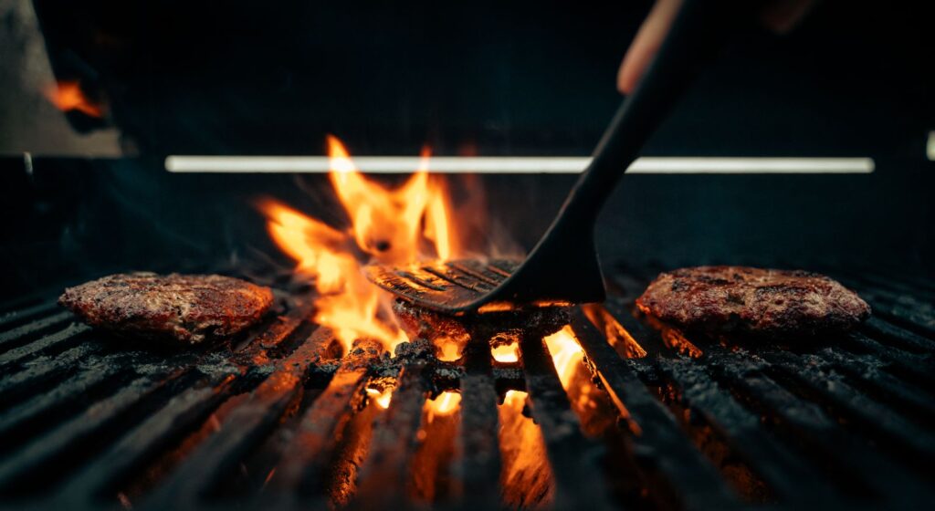 Beef patties searing over open flames on a grill, a spatula turning one