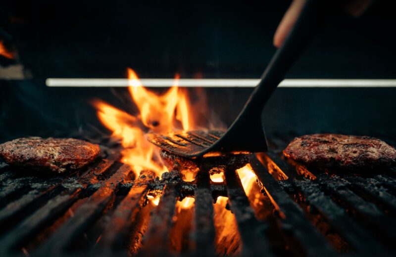 Beef patties searing over open flames on a grill, a spatula turning one