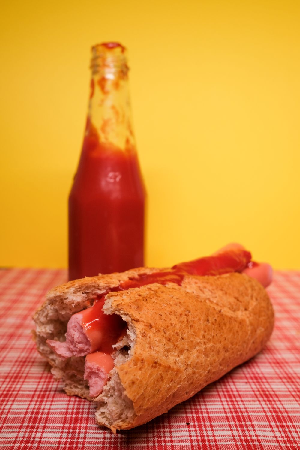 A hot dog with ketchup on a checked cloth, a glass bottle of ketchup behind it, on yellow