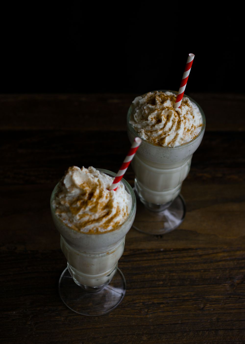 Two milkshakes topped with whipped cream and cinnamon on a rustic wooden table