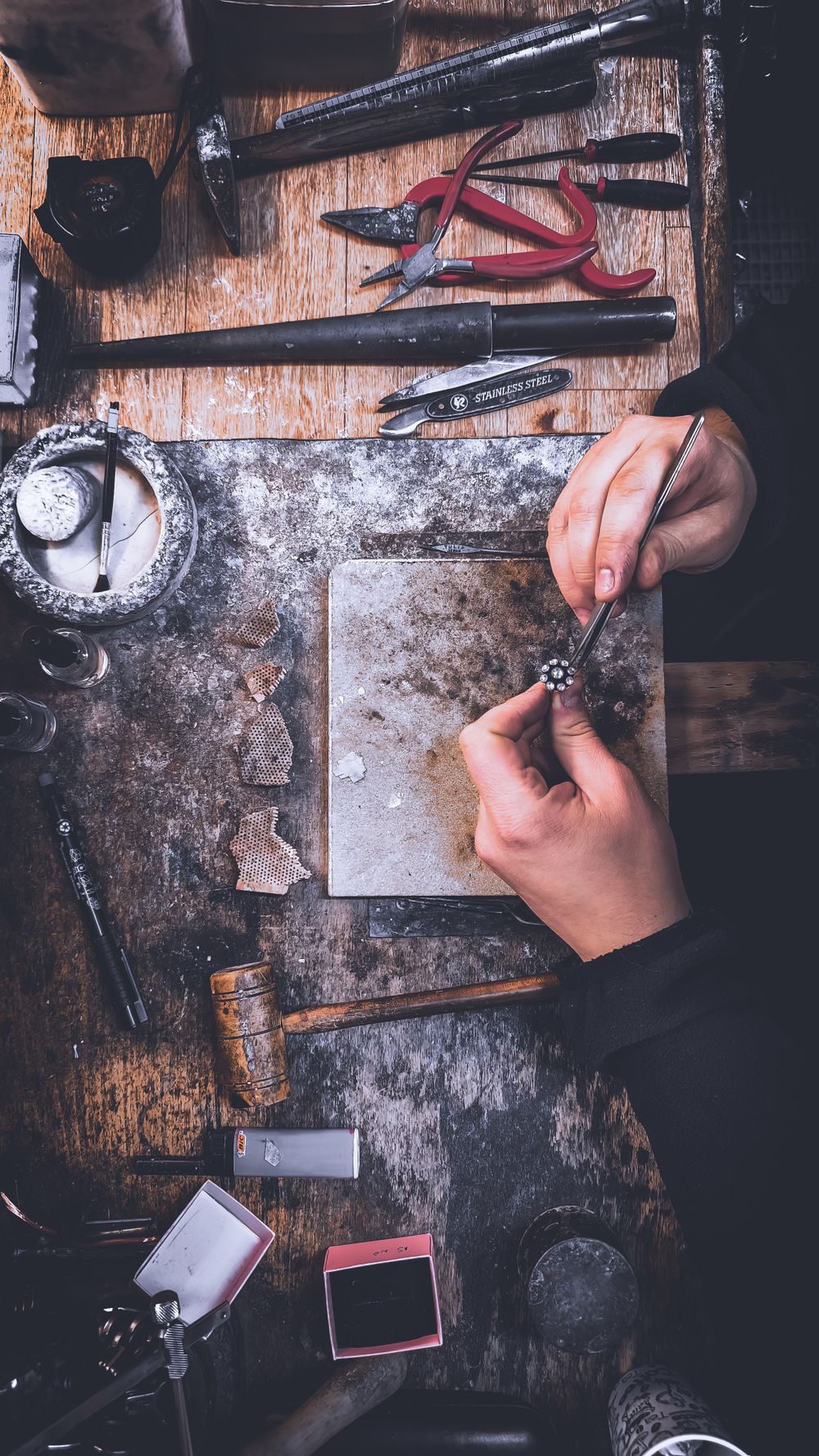 A goldsmith's hands piercing a small metal sheet with a fine tool on a worn workbench