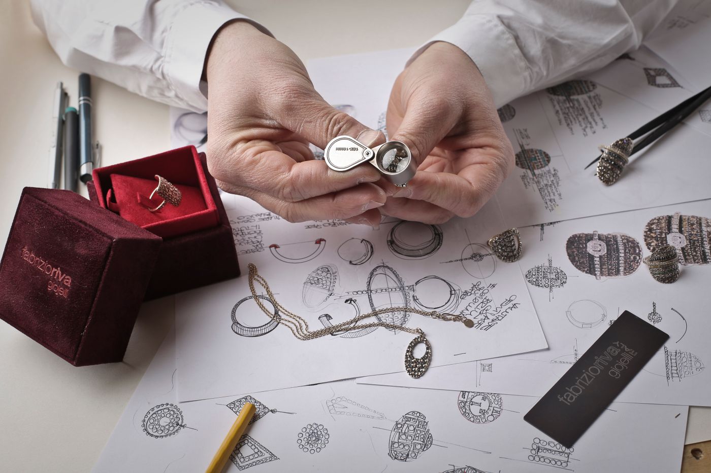 Hands checking a diamond ring with a loupe over pencil sketches of rings on the bench