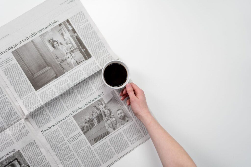 A hand holding a cup of black coffee over an open newspaper