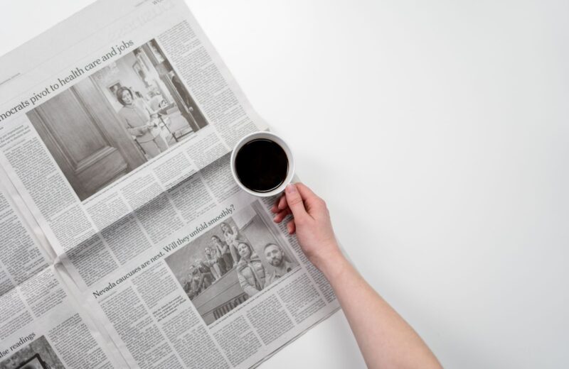 A hand holding a cup of black coffee over an open newspaper