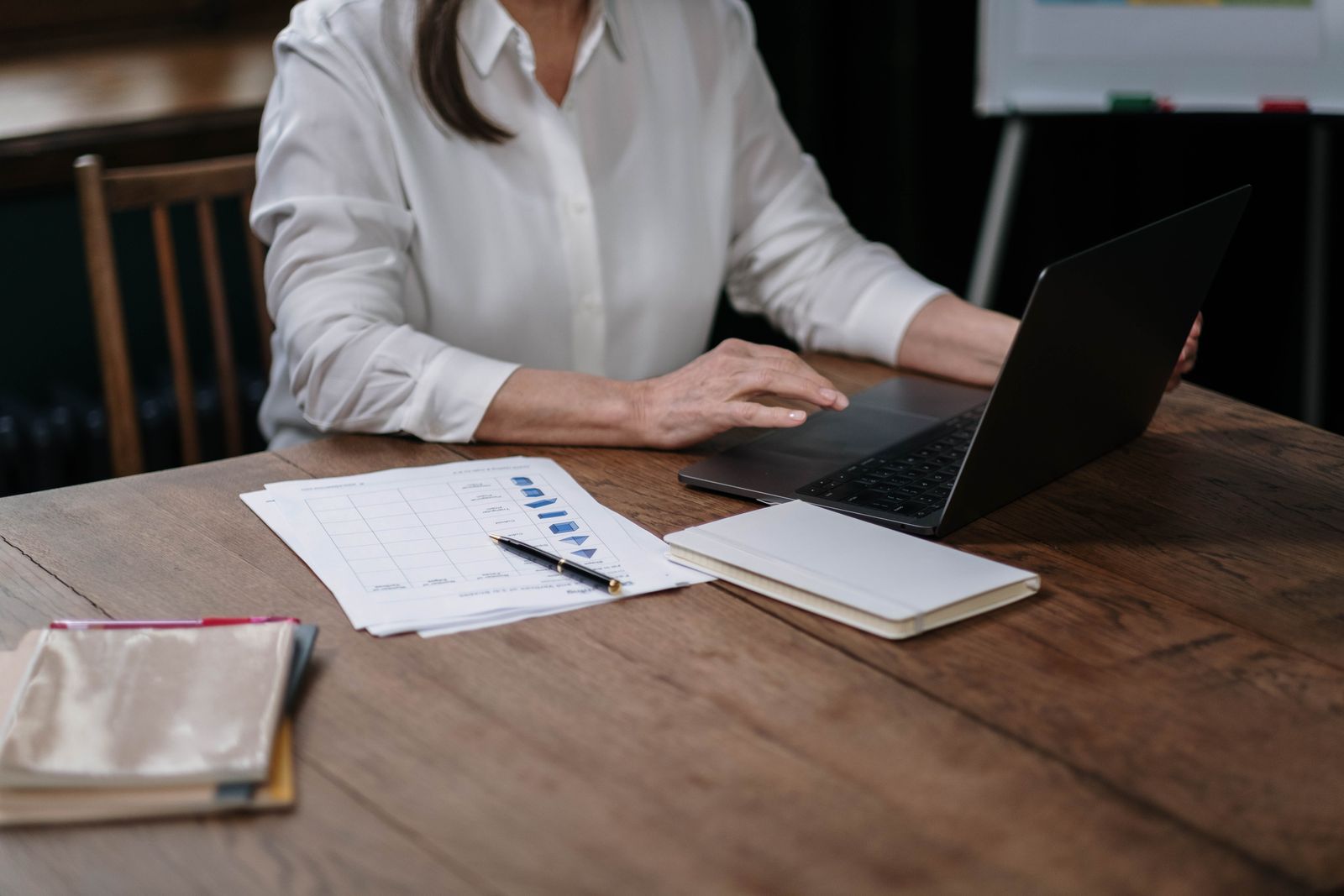 A woman working on a laptop with paper records and a notebook on a wooden desk