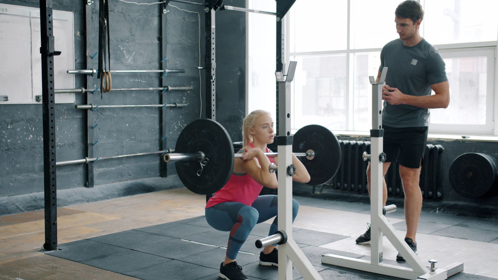 A coach watches a woman front squat a barbell in a squat rack