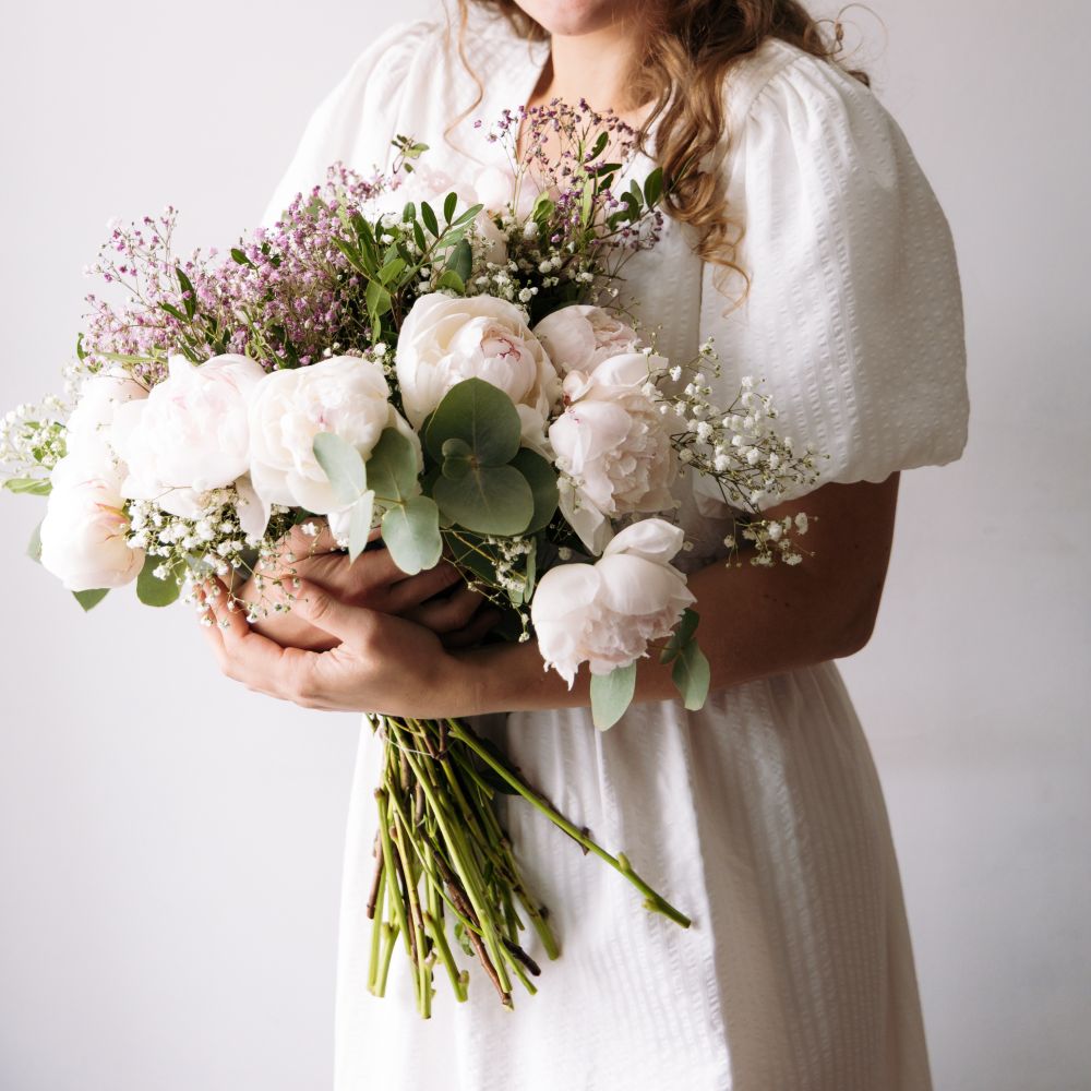 A bride in a white dress holding peonies and eucalyptus, her face out of the picture